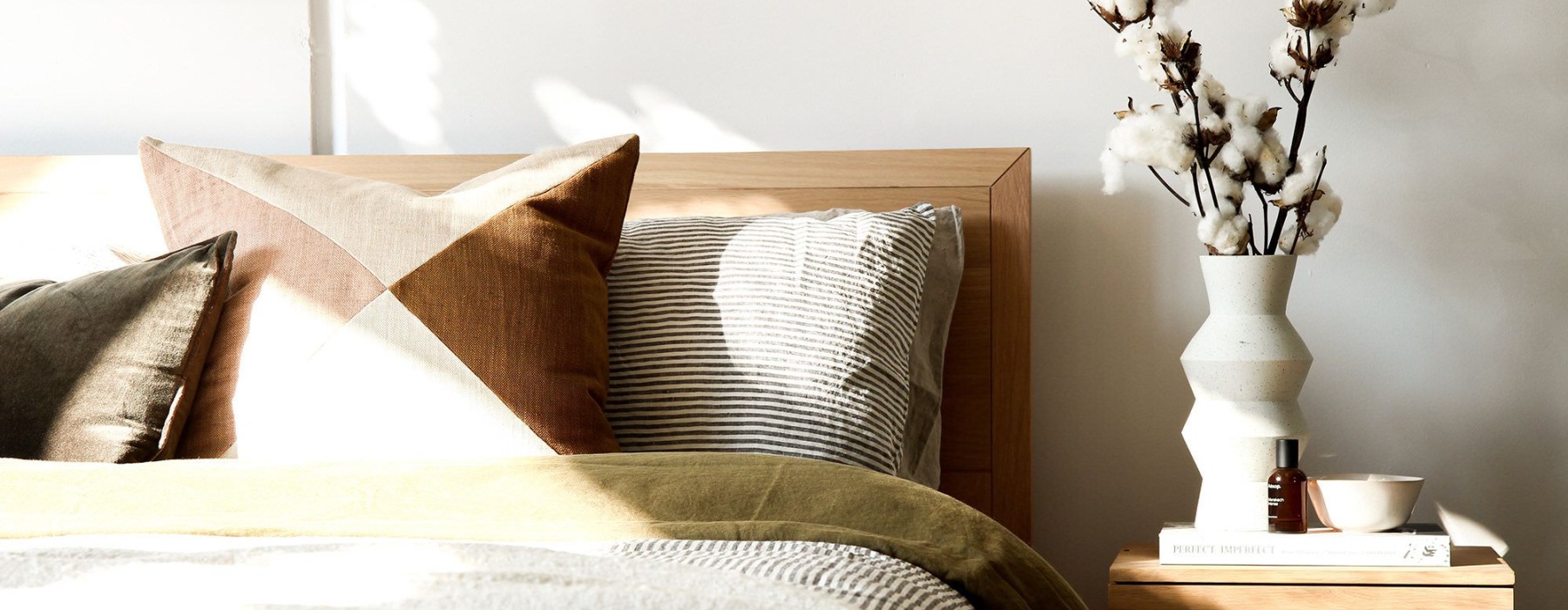 swath of sunlight shines across a well made bed and bedside table with a vase of cotton plants